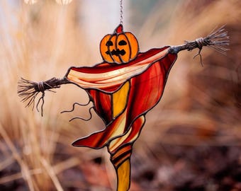 May include: A stained glass suncatcher shaped like a scarecrow with a carved orange pumpkin head and a body made of flowing red, orange, and yellow glass panels. The arms are crafted from dark metal wire, and it hangs from a thin silver chain.
