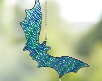 Könnte beinhalten: Ein Fledermaus-Sonnenfänger aus Buntglas mit einer strukturierten, irisierenden blau-grünen Glasoberfläche und dunklen Metalllötstellen an den Rändern. Die Fledermaus hängt an einer silberfarbenen Metallkette und hat einen kleinen Sternanhänger.