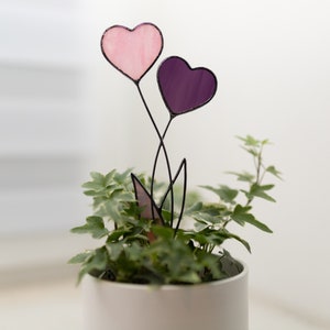 Op de afbeelding: Een roze en paarse glas-in-lood hartbloemenarrangement in een witte pot met groene, bladerige planten.