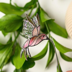 May include: A stained glass hummingbird with pink, green, and iridescent glass panels. The bird has a long, thin beak and is attached to a metal wire. The wings are spread in mid-flight, and the bird is set against a backdrop of green leaves.