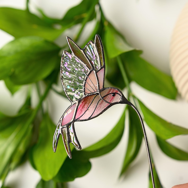 May include: A stained glass hummingbird with pink, green, and iridescent glass panels. The bird has a long, thin beak and is attached to a metal wire. The wings are spread in mid-flight, and the bird is set against a backdrop of green leaves.