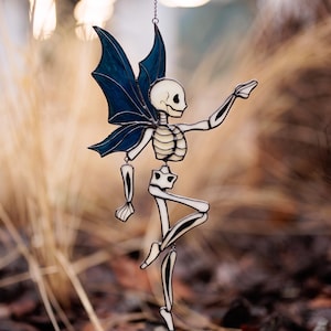 May include: A stained glass suncatcher shaped like a skeleton fairy with translucent off-white bones and dark blue wings. The figure is suspended by a thin metal chain and features black solder lines defining the skeletal structure and wing veins.