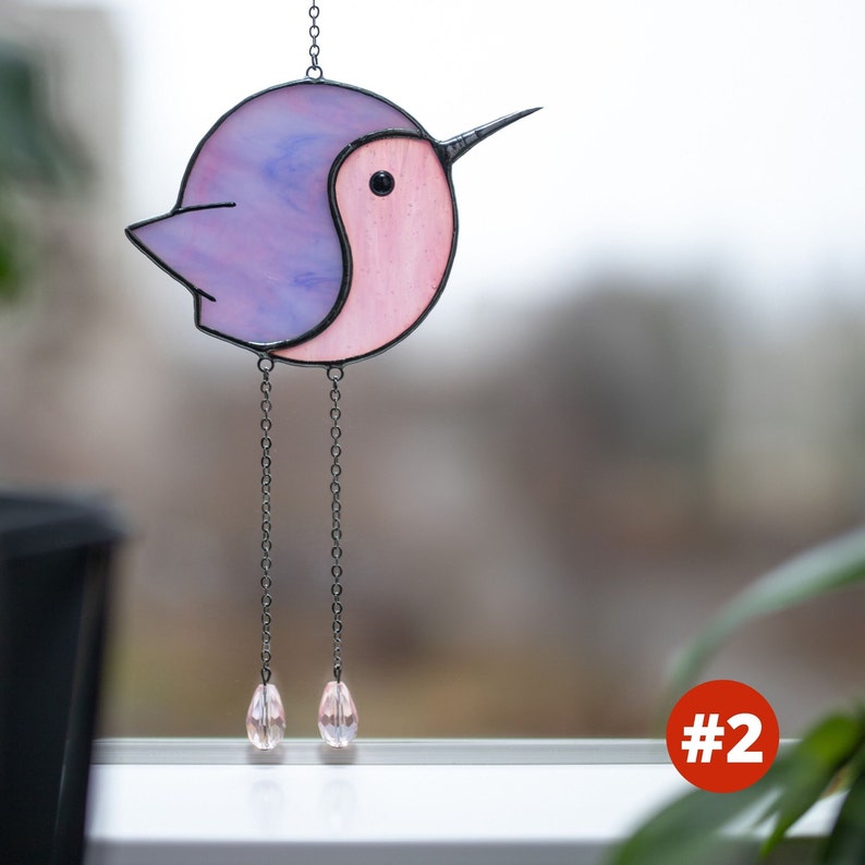 Suncatcher made with stained glass in the shape of a cartoon Hummingbird. purple hummingbird has a pink belly with long chain-legs