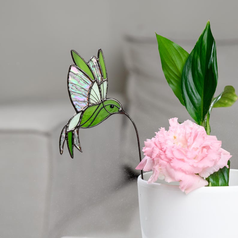 May include: A stained glass hummingbird with green and iridescent wings, a long beak, and a wire stem. The hummingbird is positioned near a pink flower and green leaves in a white pot. The background is a soft gray.