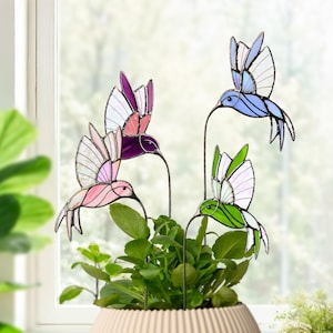 May include: Four colorful stained glass hummingbird garden stakes. The hummingbirds are in shades of pink, purple, blue, and green with white accents. They are placed in a beige pot with green plants, creating a vibrant garden decoration.
