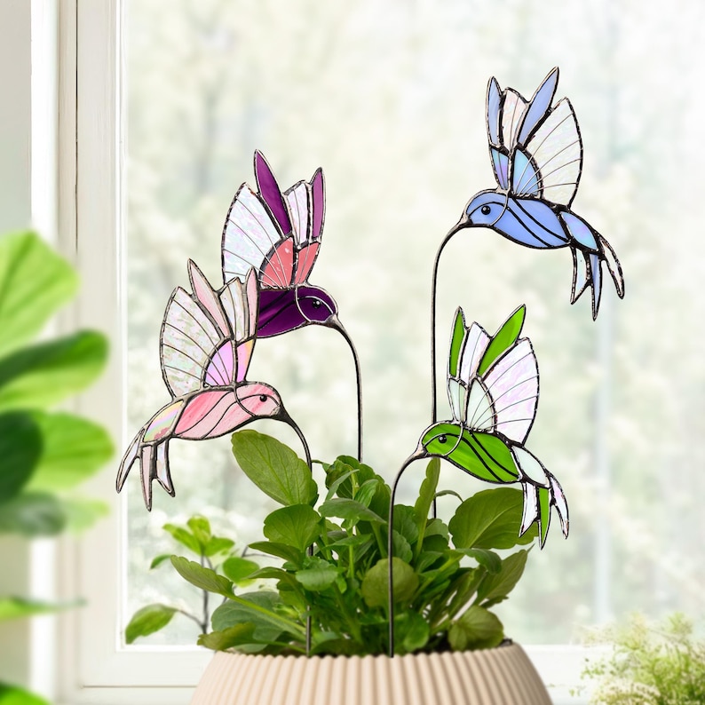 May include: Four colorful stained glass hummingbird garden stakes. The hummingbirds are in shades of pink, purple, blue, and green with white accents. They are placed in a beige pot with green plants, creating a vibrant garden decoration.