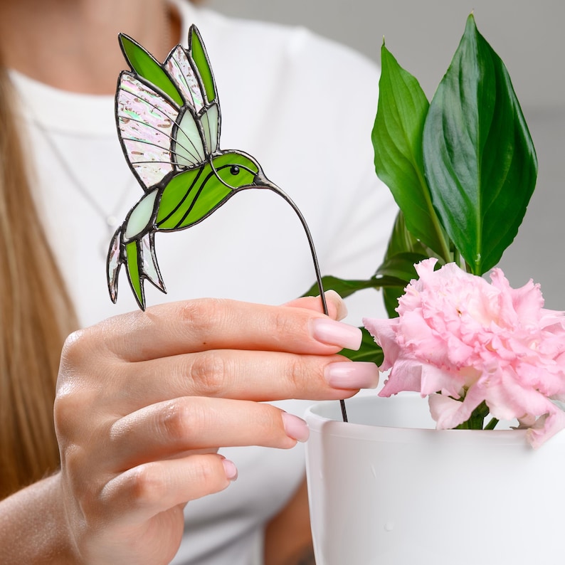 May include: A stained glass hummingbird decoration in shades of green and white, with a long, thin beak. The hummingbird is placed in a white vase with pink flowers and green leaves.