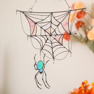 Op de afbeelding: Een glas-in-lood spinnenweb-zonnevanger met donkere metalen omlijsting en segmenten van iriserend roze en helder glas. Een bijpassende glas-in-lood spin met een glinsterend blauwgroen lichaam hangt aan een dunne draad aan het web.