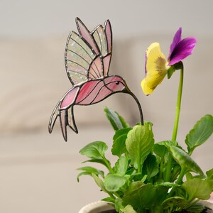 May include: A stained glass hummingbird with pink and iridescent wings hovers near a yellow and purple pansy. The hummingbird has a long, thin beak and is mounted on a metal stake. The plant has green leaves.
