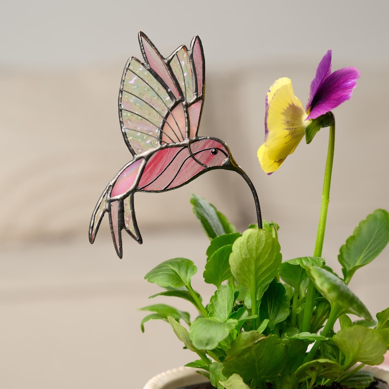 May include: A stained glass hummingbird with pink and iridescent wings hovers near a yellow and purple pansy. The hummingbird has a long, thin beak and is mounted on a metal stake. The plant has green leaves.