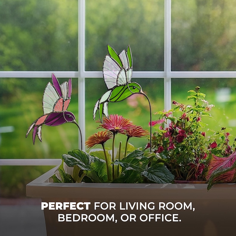 May include: Two stained glass hummingbird decorations in flight, one purple and pink, the other green and white. They are positioned in front of a window, above a planter with pink flowers and greenery. Text at the bottom reads: PERFECT FOR LIVING ROOM, BEDROOM, OR OFFICE.