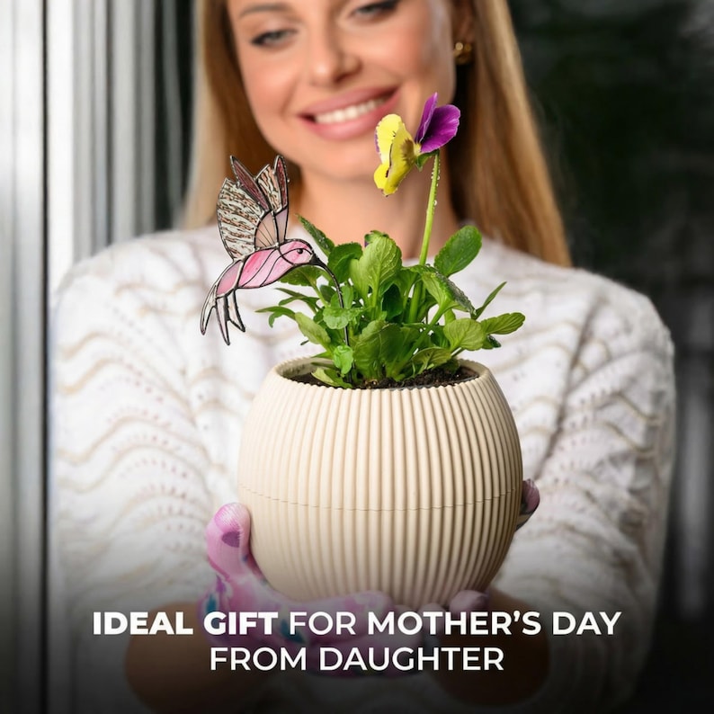 May include: A cream-colored, ribbed planter pot holds a vibrant green plant with a yellow and purple flower. A pink and brown hummingbird decoration is placed near the plant. The text at the bottom reads: IDEAL GIFT FOR MOTHER'S DAY FROM DAUGHTER.