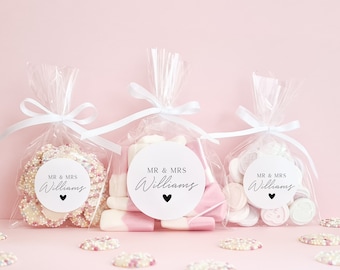 May include: Three clear cellophane bags filled with white and pink candy, tied with white ribbon. Each bag has a round white sticker with black text that reads "MR & MRS Williams" and a small black heart.