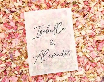 May include: A white card with the names "Isabella & Alexander" written in black calligraphy on it. The card is lying on a bed of pink and cream flower petals.