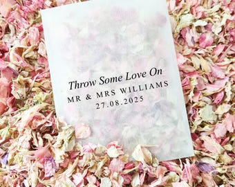 May include: A white paper bag filled with pink and white flower petals. The bag has black text that reads "Throw Some Love On MR & MRS WILLIAMS 27.08.2025".