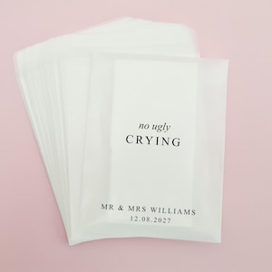 Puede incluir: Paquetes de papel blanco con el texto negro "no ugly CRYING". Debajo, los nombres "MR & MRS WILLIAMS" y la fecha "12.08.2027". Apilados sobre un fondo rosa claro.