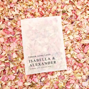 Może przedstawiać: Biała papierowa torebka z napisem "THROW SOME LOVE" i imionami "ISABELLA & ALEXANDER" z datą "SUNDAY 30TH AUGUST 2026" wydrukowanymi na czarno. Torebka spoczywa na podłożu z różowych i kremowych płatków kwiatów.