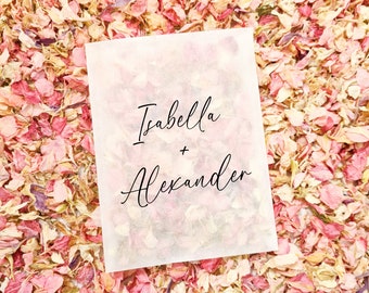 May include: A white paper bag with the text "Isabella + Alexander" written in black cursive script. The bag is surrounded by pink and white flower petals.
