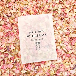 May include: A white packet with the text "MR & MRS WILLIAMS" and the date "28.08.2027" printed in black. A small bow illustration is below the date. The packet is surrounded by pink and peach flower petals.