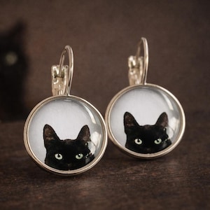 May include: A pair of round silver-toned drop earrings featuring a glass cabochon insert. Each earring displays a detailed photographic print of a black cat with bright green eyes against a white background, suspended from a silver-toned leverback.