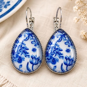 Può includere: Un paio di orecchini a forma di goccia con un motivo floreale blu e bianco sotto una cupola di vetro. Gli orecchini sono montati su una cornice in metallo color argento con chiusure a leva, mostrando un design botanico dettagliato su sfondo bianco.