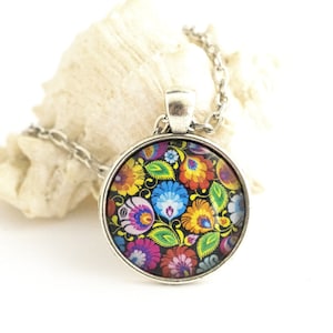 Puede incluir: Un collar con colgante redondo de plata que presenta un vibrante patr&oacute;n floral multicolor sobre un fondo negro. El dise&ntilde;o incluye intrincados motivos de flores y hojas en rojo, azul, amarillo y verde dentro de un marco de metal plateado pulido.