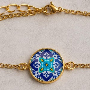 May include: A gold-toned chain bracelet featuring a central circular charm with a vibrant blue, teal, and white geometric floral pattern. The bracelet includes a delicate link chain with a lobster claw clasp and an adjustable extender chain.