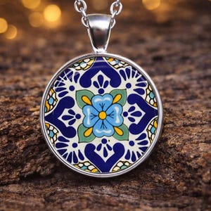 May include: A silver pendant necklace featuring a circular charm with a vibrant floral pattern in shades of blue, green, yellow, and white. The design displays a central light blue flower surrounded by intricate dark blue and yellow geometric motifs.
