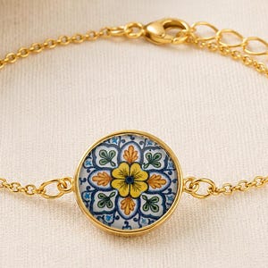 May include: A gold-tone chain bracelet featuring a central circular charm with a white, yellow, and blue floral tile pattern. The bracelet includes a delicate link chain, a lobster claw clasp, and an adjustable extender chain with a small gold bead.