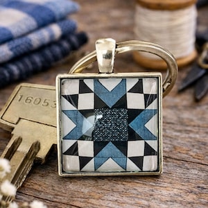 May include: A square metal keychain featuring a geometric star pattern in shades of blue, black, and white. The center of the design is a dark, textured square surrounded by alternating triangles and squares, all encased in a silver-toned frame.