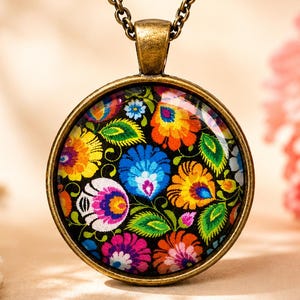 Puede incluir: Un collar con colgante redondo que presenta un engaste de metal en tono bronce y un caboch&oacute;n de vidrio. El inserto muestra un patr&oacute;n floral vibrante y multicolor con p&eacute;talos rojos, amarillos, azules y verdes sobre fondo negro.