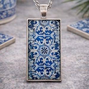 May include: A rectangular pendant necklace featuring a silver-toned metal frame and a blue and white patterned insert. The design displays a repeating floral and geometric motif reminiscent of traditional tile work, suspended from a silver chain.
