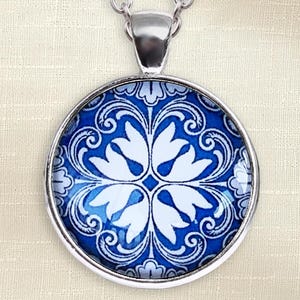 Puede incluir: Un colgante circular de plata con un vibrante patr&oacute;n floral en azul y blanco. El dise&ntilde;o consiste en p&eacute;talos blancos sim&eacute;tricos dispuestos en forma de cruz sobre un fondo azul intenso con detalles intrincados de volutas blancas.