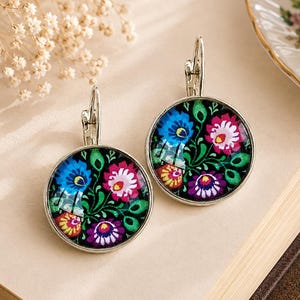 May include: A pair of round silver-toned leverback earrings featuring a vibrant floral pattern on a black background. The design includes stylized blue, pink, purple, and yellow flowers with green leaves encased under a smooth glass dome surface.