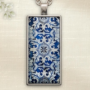 May include: Silver pendant necklace with a rectangular pendant displaying a blue and white tile pattern. The pendant features a symmetrical design of floral and geometric shapes. The necklace has a silver chain.