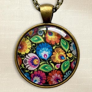 Puede incluir: Un colgante redondo con un marco de metal en tono bronce y un cabuj&oacute;n de cristal. El inserto muestra un patr&oacute;n floral multicolor vibrante con flores estilizadas en tonos rosa, azul, naranja y amarillo sobre un fondo negro.