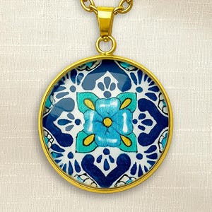 Peut inclure: Un collier pendentif doré avec un pendentif rond. Le pendentif présente un motif floral bleu et blanc avec des détails turquoise et jaunes. La chaîne est une chaîne à maillons dorés.