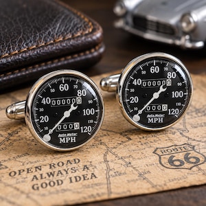 Op de afbeelding: Een paar ronde zilverkleurige manchetknopen met een zwarte snelheidsmeter wijzerplaat, witte markeringen, een sleutelvormige naald en de tekst Auto Meter MPH. De meter toont getallen van 0 tot 120 en een digitale kilometerteller met 000000.