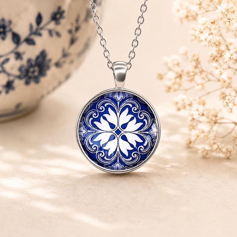 Puede incluir: Un collar de tono plateado con un colgante circular que presenta un patr&oacute;n floral en azul y blanco. El colgante cuelga de una delicada cadena plateada, mostrando un dise&ntilde;o sim&eacute;trico con p&eacute;talos blancos intrincados sobre un fondo azul intenso.