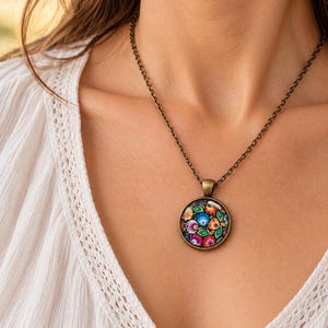 Puede incluir: Un collar con colgante redondo que presenta un patr&oacute;n floral vibrante y multicolor dentro de un marco de metal en tono bronce antiguo, suspendido de una fina cadena met&aacute;lica a juego sobre una prenda de tela blanca texturizada.