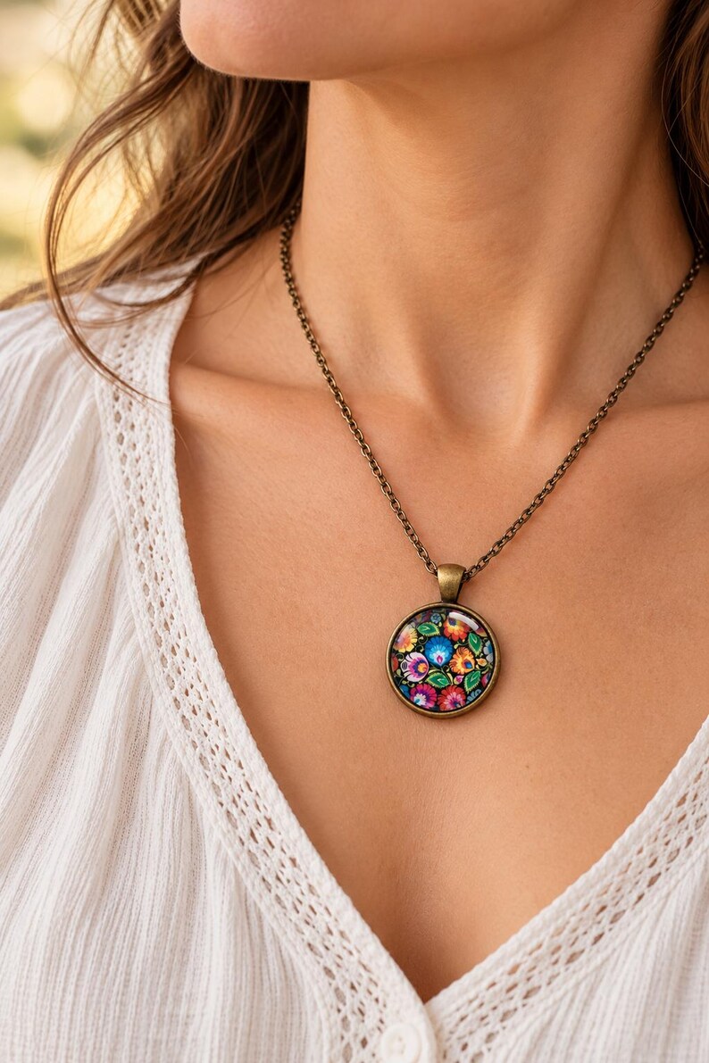 Puede incluir: Un collar con colgante redondo que presenta un patr&oacute;n floral vibrante y multicolor dentro de un marco de metal en tono bronce antiguo, suspendido de una fina cadena met&aacute;lica a juego sobre una prenda de tela blanca texturizada.