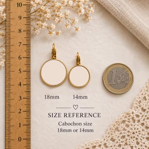 Puede incluir: Dos pendientes circulares dorados con centros color crema en blanco. Una regla de madera muestra que el pendiente m&aacute;s grande mide 18 mm y el m&aacute;s peque&ntilde;o 14 mm. Texto: 18mm, 14mm, SIZE REFERENCE, Cabochon size 18mm or 14mm. Una moneda de un euro sirve de escala.