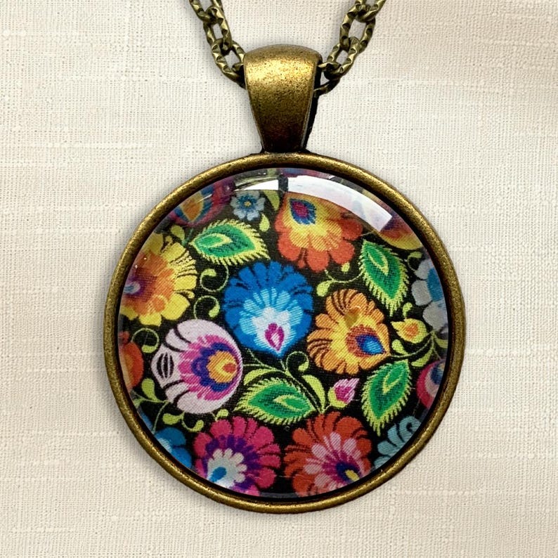 Puede incluir: Un colgante redondo con un marco de metal en tono bronce y un cabuj&oacute;n de cristal. El inserto muestra un patr&oacute;n floral multicolor vibrante con flores estilizadas en tonos rosa, azul, naranja y amarillo sobre un fondo negro.