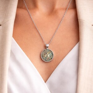 May include: A silver-toned necklace featuring a circular pendant with a multifaceted, iridescent geometric inlay. The pendant hangs from a delicate silver-toned chain, showcasing a reflective surface with shifting hues of green, blue, and gold.