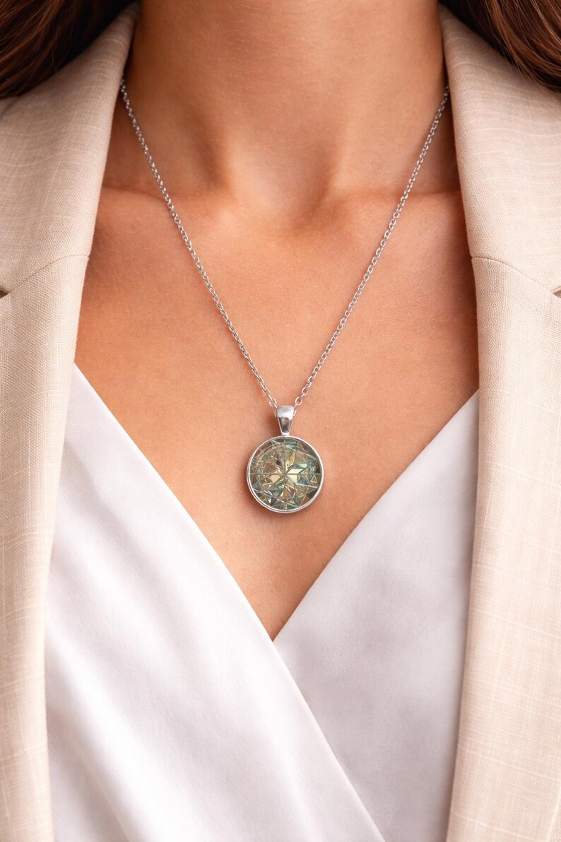 May include: A silver-toned necklace featuring a circular pendant with a multifaceted, iridescent geometric inlay. The pendant hangs from a delicate silver-toned chain, showcasing a reflective surface with shifting hues of green, blue, and gold.