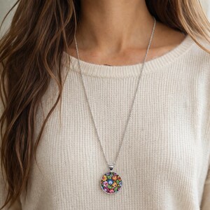 Puede incluir: Un collar de tono plateado con un colgante circular que presenta un patr&oacute;n floral multicolor y vibrante sobre un fondo negro. El colgante cuelga de una cadena fina y delicada de tono plateado que descansa sobre un su&eacute;ter de punto color crema.