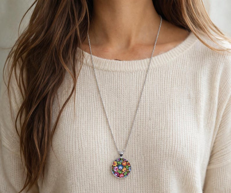 Puede incluir: Un collar de tono plateado con un colgante circular que presenta un patr&oacute;n floral multicolor y vibrante sobre un fondo negro. El colgante cuelga de una cadena fina y delicada de tono plateado que descansa sobre un su&eacute;ter de punto color crema.