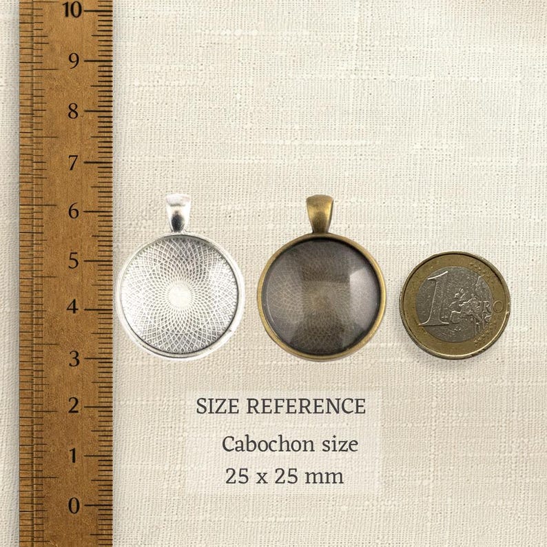Puede incluir: Dos bases para colgante redondas de metal, una plateada y otra bronce, junto a una regla de madera y una moneda de un euro. El texto dice SIZE REFERENCE Cabochon size 25 x 25 mm. Las bases cuentan con un engaste circular.