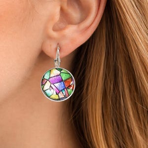 Puede incluir: Un pendiente redondo de plata con cierre de palanca que presenta un patr&oacute;n de mosaico multicolor con segmentos en verde, morado, naranja, azul y amarillo. El dise&ntilde;o circular est&aacute; enmarcado en plata pulida, creando un efecto de vidriera.