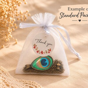 Puede incluir: Una bolsa de organza blanca con cord&oacute;n que contiene un collar con un colgante de ojo de pluma de pavo real. Una etiqueta redonda blanca unida al cord&oacute;n presenta el texto Thank you y un peque&ntilde;o gr&aacute;fico de corona floral en rojo y verde.